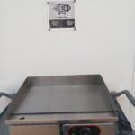 Front view of a 55cm commercial griddle with a flat stainless steel cooking plate, partially visible temperature controls, and an easy-access oil waste collection drawer for efficient commercial kitchen use.