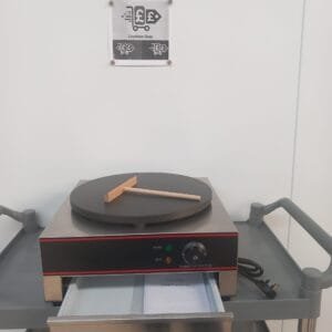 Front view of a commercial 400mm crepe maker with Teflon-coated plate, crepe spreader on the cooking surface, and drawer below open to display the user manual, designed for easy operation in commercial kitchens.