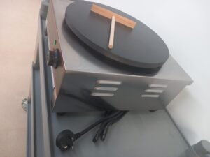 Slight aerial view from the right of a commercial 400mm crepe maker with Teflon-coated plate and crepe spreader, showing the plug in closer detail for a more comprehensive look at the appliance setup.